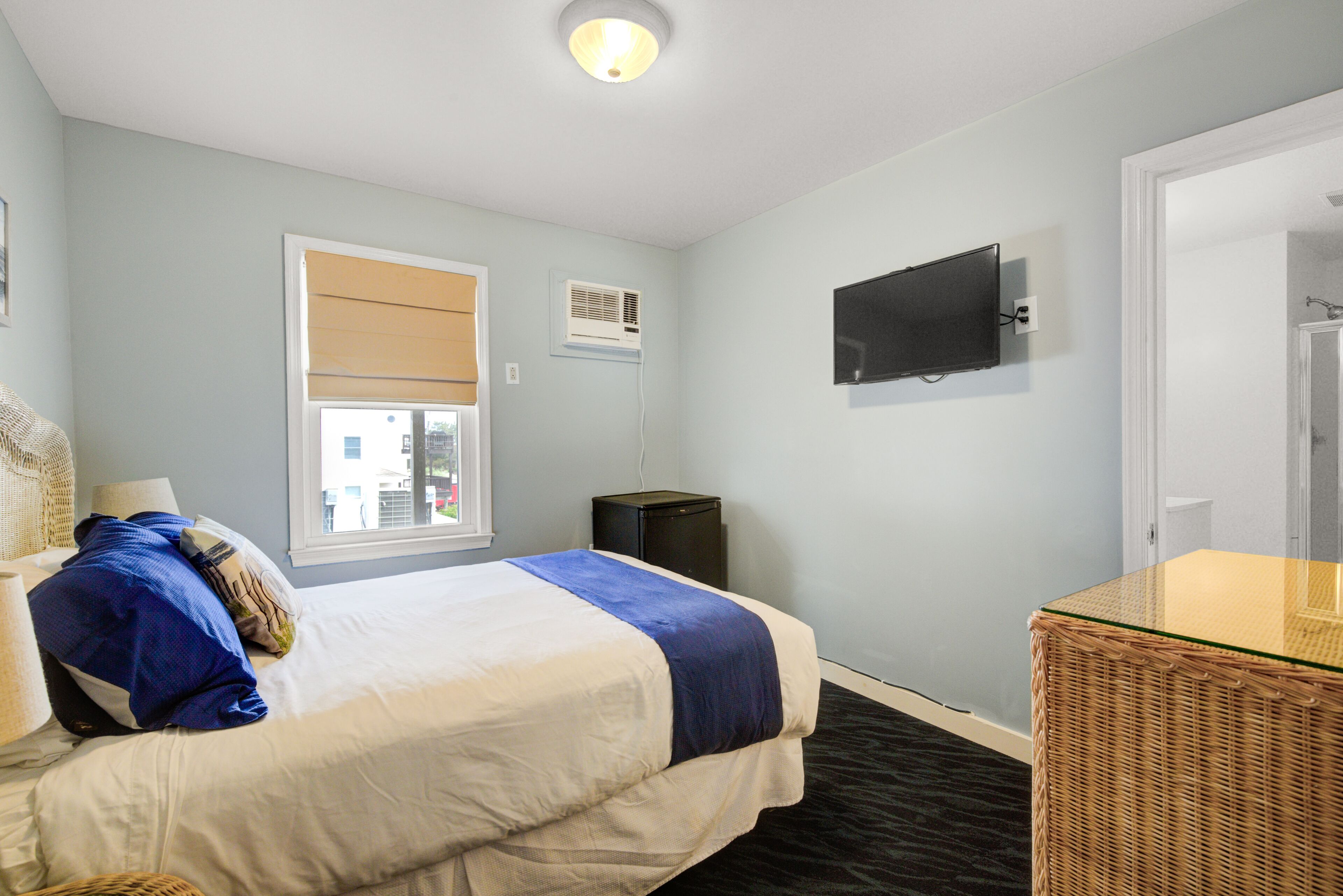A hotel room with a bed, a television, a window with blinds, a small table, and a wicker chair.