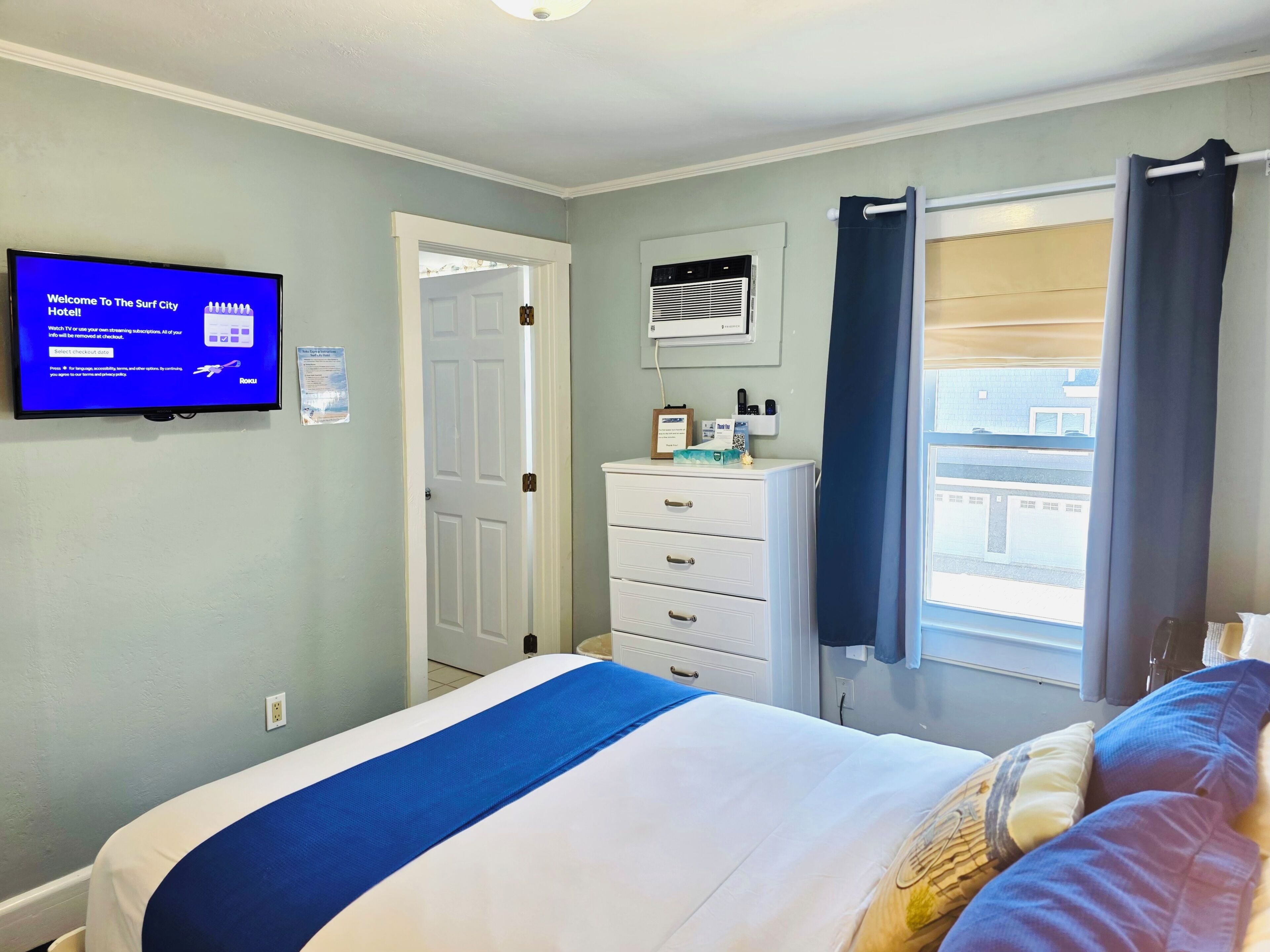 A hotel room with a bed, a dresser, a television displaying a welcome message, a door, and a window with curtains.