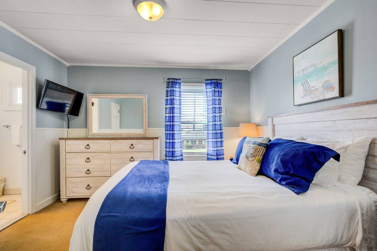 A bedroom with a bed, a dresser, a television, a window with curtains, and a painting on the wall.