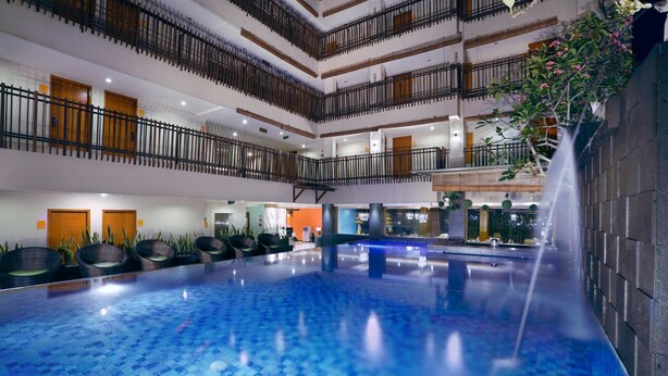 A multi-level indoor swimming pool with a waterfall feature, surrounded by seating areas and a modern architectural design. - Sun Royal Hotel (Kuta)
