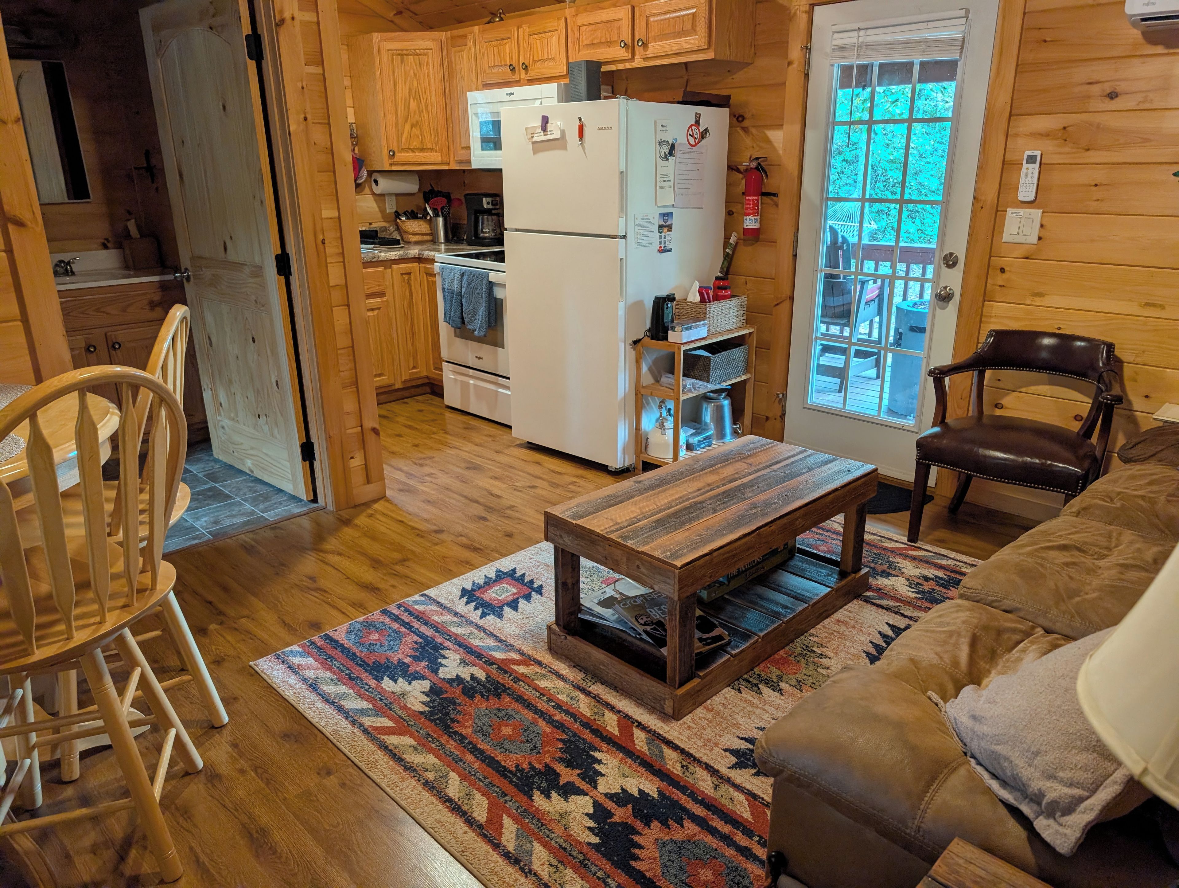 A cozy cabin interior with a wooden dining table, a sofa, and a kitchen area with a refrigerator and wooden cabinets.