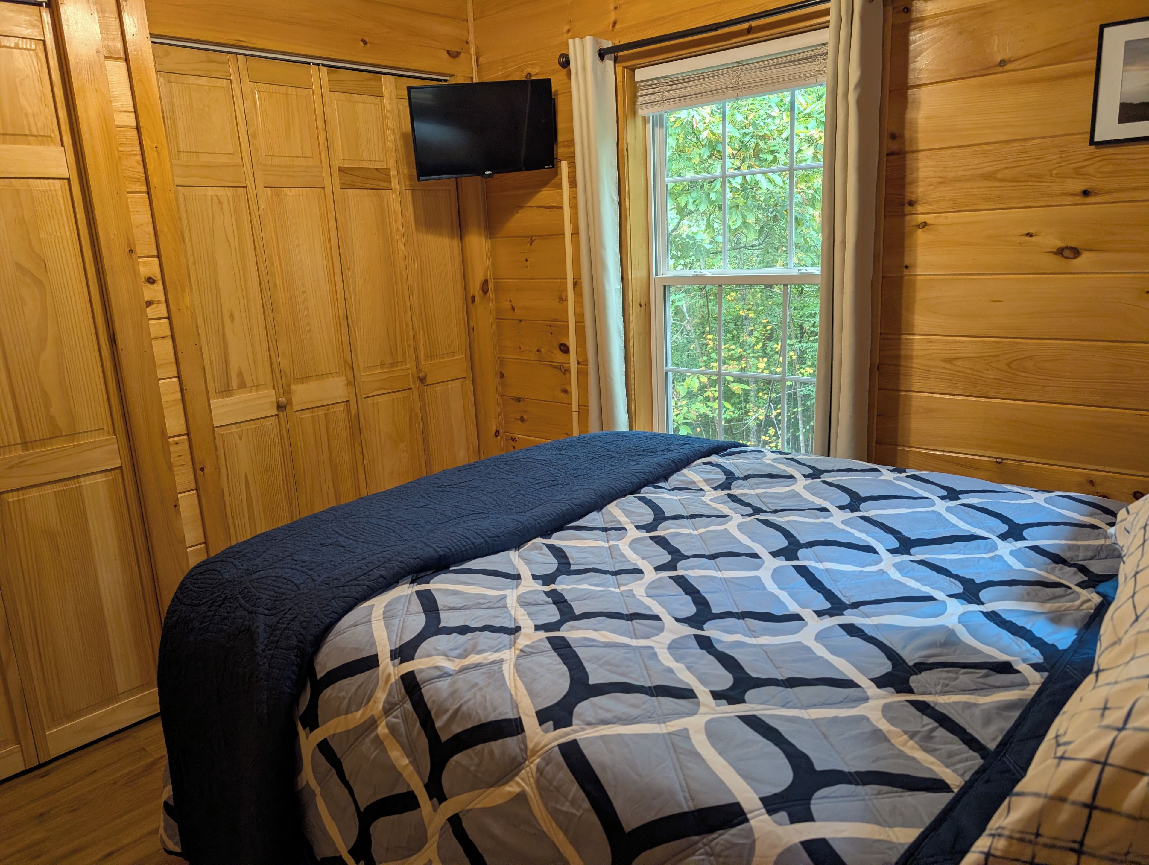 A wooden room with a bed, a television mounted on the wall, and a window with a view of trees.