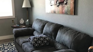 Un salon avec un canapé en cuir foncé, un tapis à motifs, une œuvre d’art représentant une voiture ancienne accrochée au mur, et une table d’appoint avec une lampe.