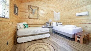 A wooden cabin bedroom with two beds, a small table, a lamp, and a wall-mounted air conditioner. - 1bd rm, 1 bth, hotel style cabin w/ Mini fridge, micro, & tv, at Kick Back Ranch (Ramer)