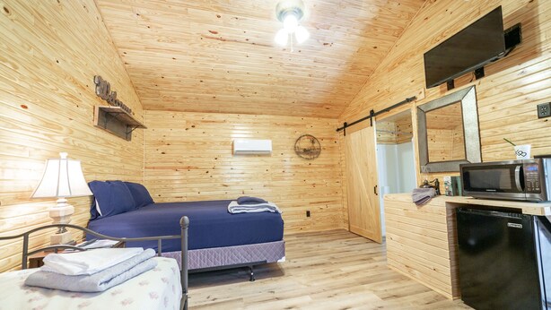 A compact wooden cabin room with a bed, a desk with a lamp, a microwave, and a small refrigerator. - 1bd rm, 1 bth, hotel style cabin w/ Mini fridge, micro, & tv, at Kick Back Ranch (Ramer)