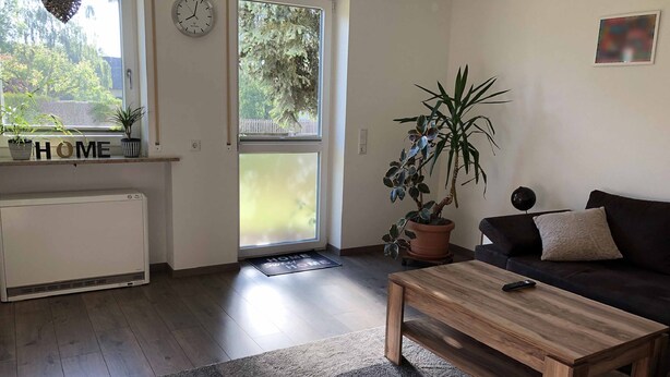 A living room with a sofa, coffee table, and a potted plant. There is a window with a view of trees and a clock on the wall. - Burgauer Fewo (Burgau)