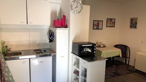 A compact kitchen with a refrigerator, a microwave, and a small stove. There is a clock on the wall and a chair in the dining area. - Burgauer Fewo (Burgau)