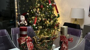 Árvore de Natal com enfeites e luzes, um boneco do Papai Noel e presentes embrulhados em uma mesa de vidro em um quarto com cadeiras roxas e um abajur.