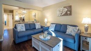 Una sala de estar con un sofá azul, una mesa de centro con una bandeja decorativa, un ventilador de techo y una cocina visible al fondo.