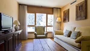 A living room with a wooden coffee table, a sofa with striped cushions, a striped armchair, a television on a cabinet, and large windows with curtains. - Solei - Edificio Bonaigua (Baqueira)