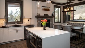 Une cuisine moderne avec des meubles blancs, un îlot central et une cheminée. Des luminaires suspendus sont placés au-dessus de l’îlot, et une grande fenêtre laisse entrer la lumière naturelle.
