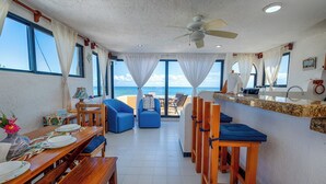Amplia habitación con un gran ventanal con vistas al mar, ventilador de techo y zona de comedor con muebles de madera.