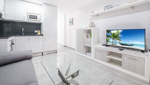 Un salon moderne doté d’un téléviseur à écran plat, d’un canapé gris et d’un coin cuisine avec des meubles blancs et un four à micro-ondes.
