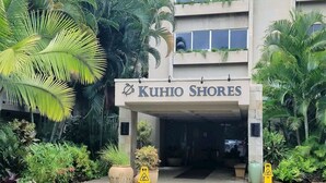 Ingången till ett hotell som heter ”Kuhio Shores”, med en skylt ovanför ingången, omgiven av frodig grönska och palmer.
