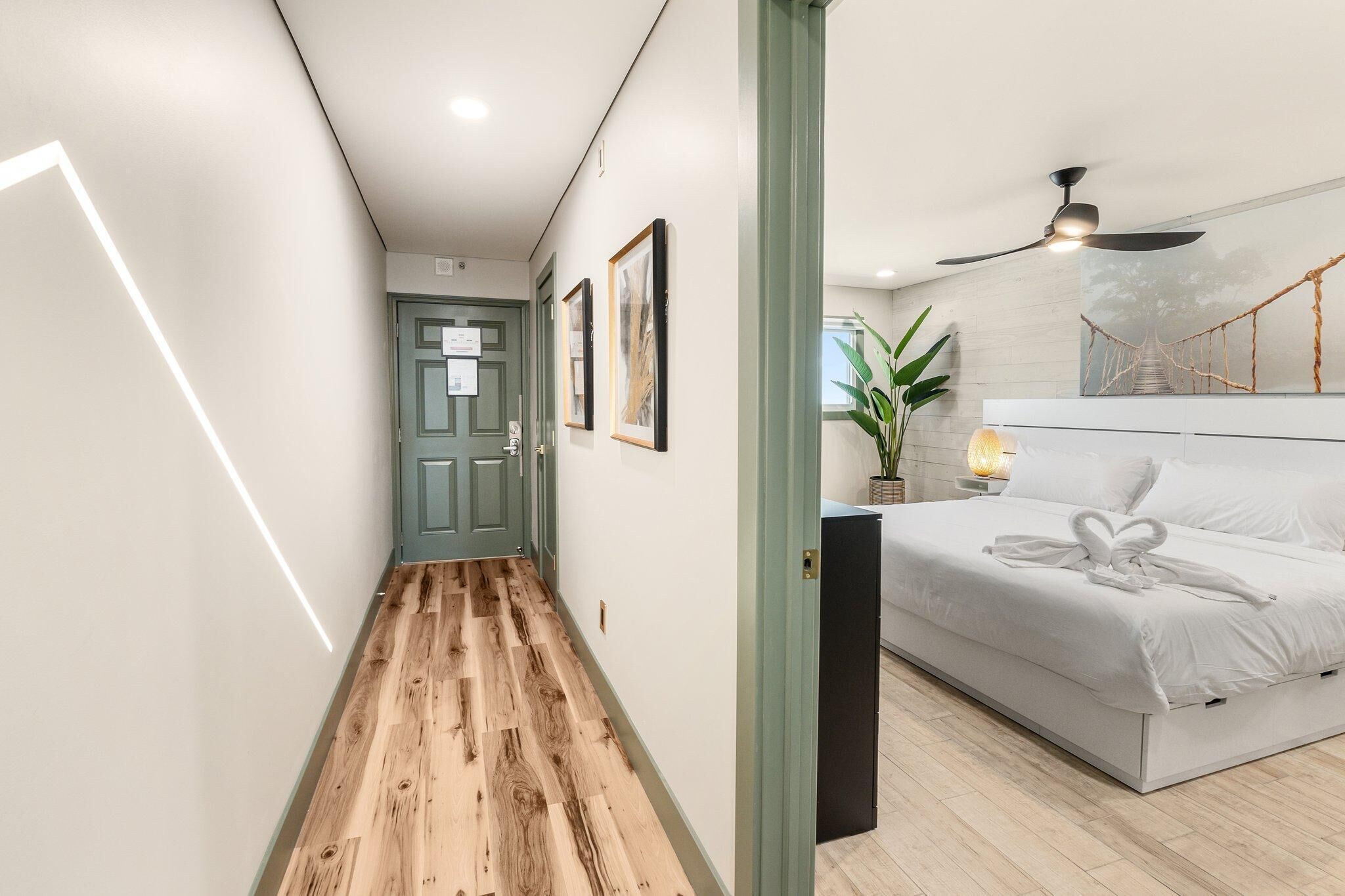 A modern hotel room with a wooden floor hallway, a bed with a heart-shaped towel, a ceiling fan, and a wall-mounted plant.