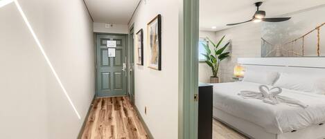 A modern hotel room with a wooden floor hallway, a bed with a heart-shaped towel, a ceiling fan, and a wall-mounted plant.