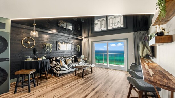 A modern living room with a wooden floor, a black sofa with patterned cushions, a dining area with a table and chairs, and a large window with a view of the sea. - The Summit 1107 (Panama City Beach)
