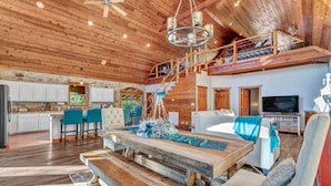 A spacious living area with a wooden ceiling, a dining table, and a TV in the background.