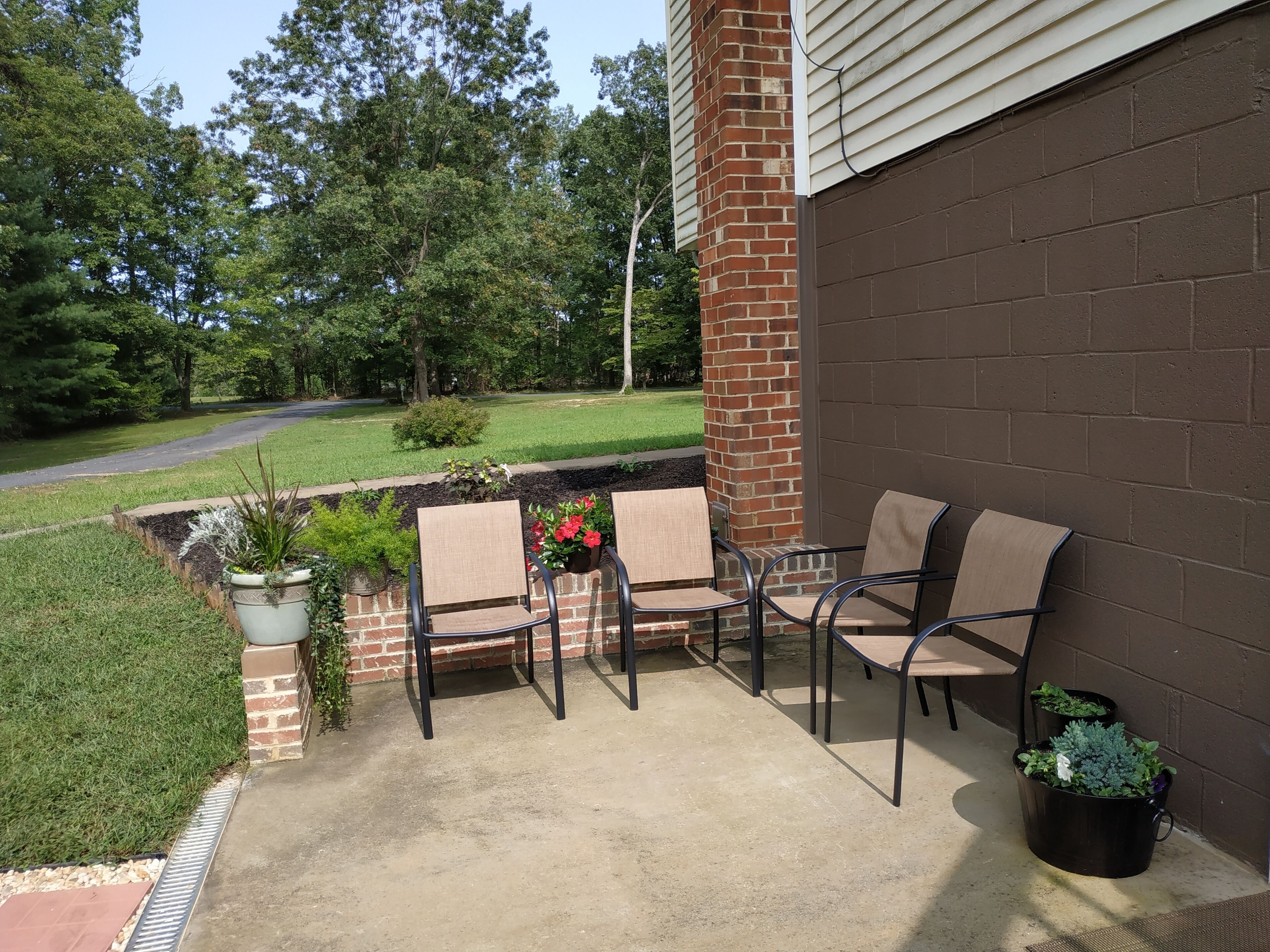 A patio with four chairs and two potted plants, a brick wall, and a grassy area with trees in the background.