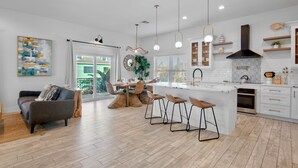 A modern kitchen with white cabinetry, a central island with bar stools, pendant lighting, and a large window. - Paradise Bound! Backyard Bliss with Sun Shelf, BBQ and Coastal Decor (Anna Maria)