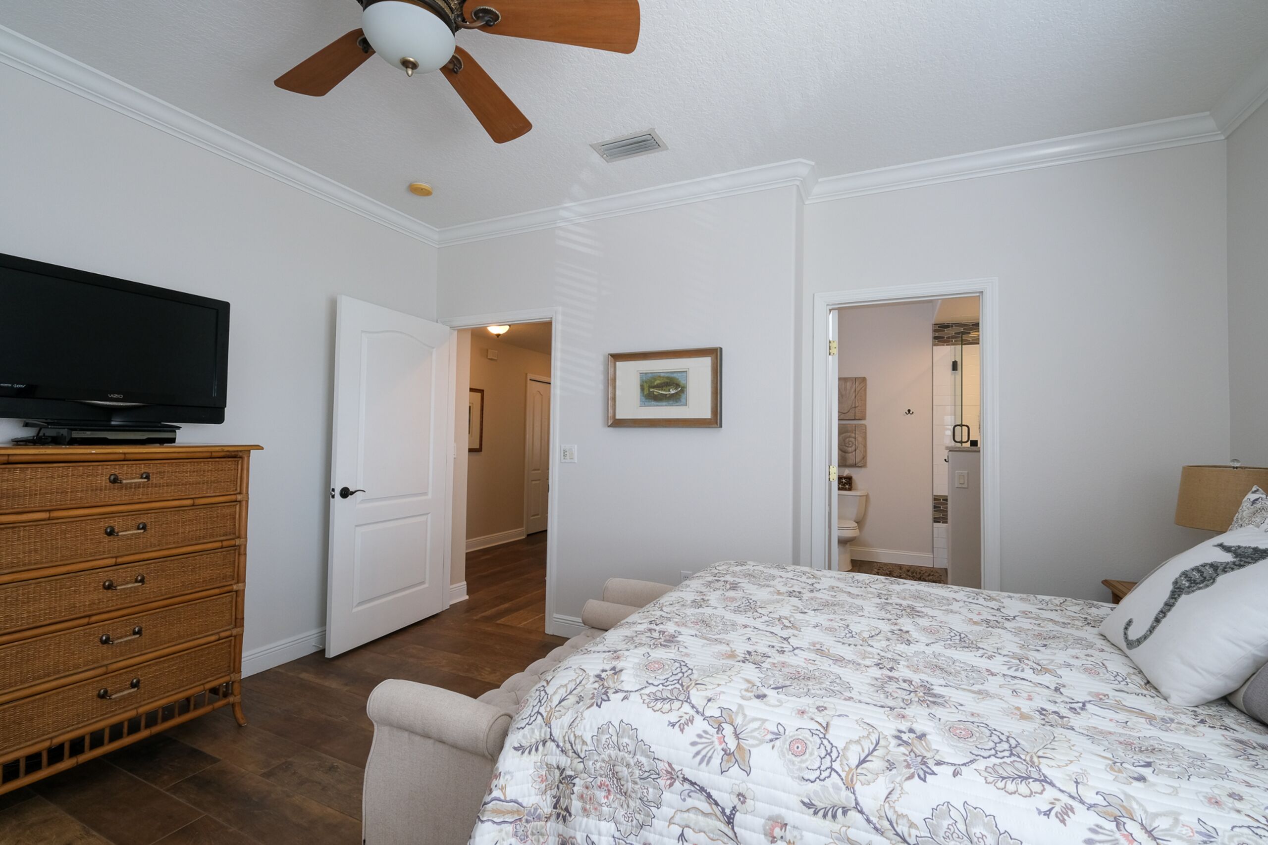A bedroom with a bed, a television on a dresser, a ceiling fan, and a door leading to another room.