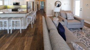 A modern kitchen with white cabinets, a central island with bar stools, and a dining area with a gray sofa and patterned rug.