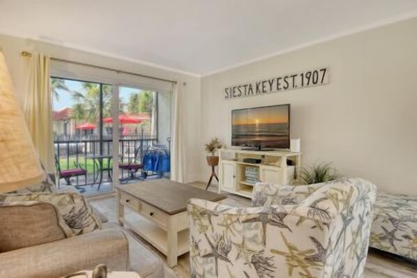 A living room with a sofa, a coffee table, and a TV on a stand. There is a sliding glass door leading to an outdoor area with chairs and umbrellas. - Condo# 119 Beautifull updated Condo Siesta Key (Sarasota)