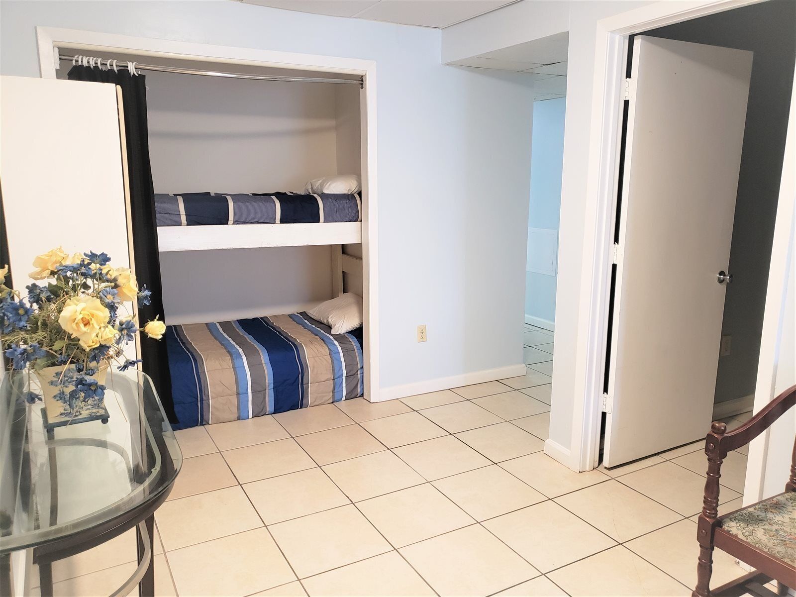 Quarto pequeno com beliche, mesa de vidro com um vaso de flores e porta que dá para outro quarto.