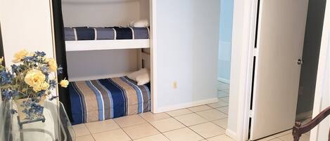 Quarto pequeno com beliche, mesa de vidro com um vaso de flores e porta que dá para outro quarto.