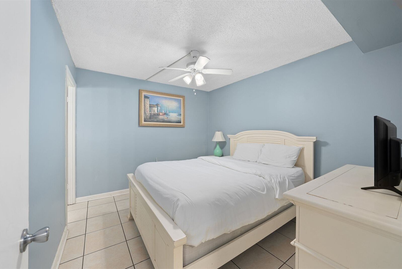 A bedroom with a bed, a ceiling fan, a bedside table, a lamp, and a TV.
