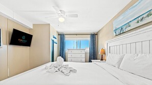 A hotel room with a bed, a ceiling fan, a TV, a dresser, and a painting of a beach scene. - The Summit 1011 (Panama City Beach)