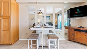 A dining area with a white table, two cushioned stools, and a ceiling fan. A TV is mounted on the wall to the right. - The Summit 1011 (Panama City Beach)