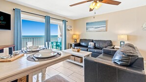 A modern living room with a dining area, a ceiling fan, a view of the ocean, and a sign for "Quest Vacation Rentals". - The Summit 1011 (Panama City Beach)