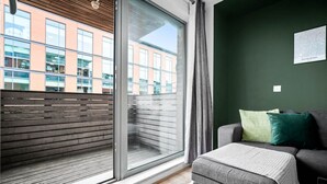 Una camera moderna con una porta a vetri scorrevoli che dà su un balcone esterno, un divano grigio con cuscini e una parete verde con un quadro incorniciato.