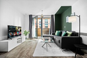 Una sala de estar moderna con un televisor de pantalla plana, un sofá, una mesa de centro de vidrio y vistas a un edificio a través de una puerta de cristal.
