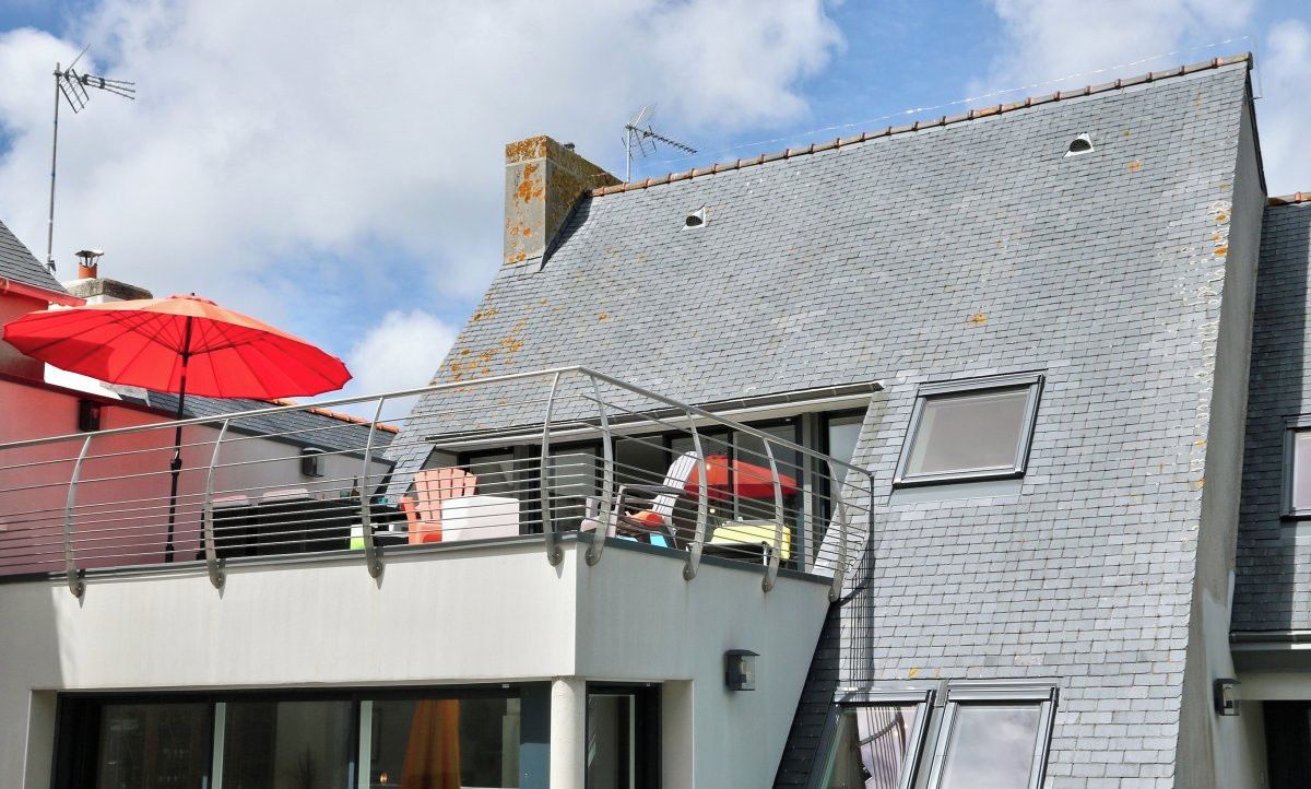 A residential building with a grey tiled roof, a balcony with a red umbrella, and a visible window.