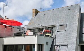 Un immeuble résidentiel avec un toit recouvert de tuiles grises, un balcon agrémenté d’un parasol rouge et une fenêtre bien visible.