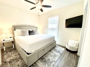 A bedroom with a bed, a nightstand, a ceiling fan, a window with blinds, and a wall-mounted TV.