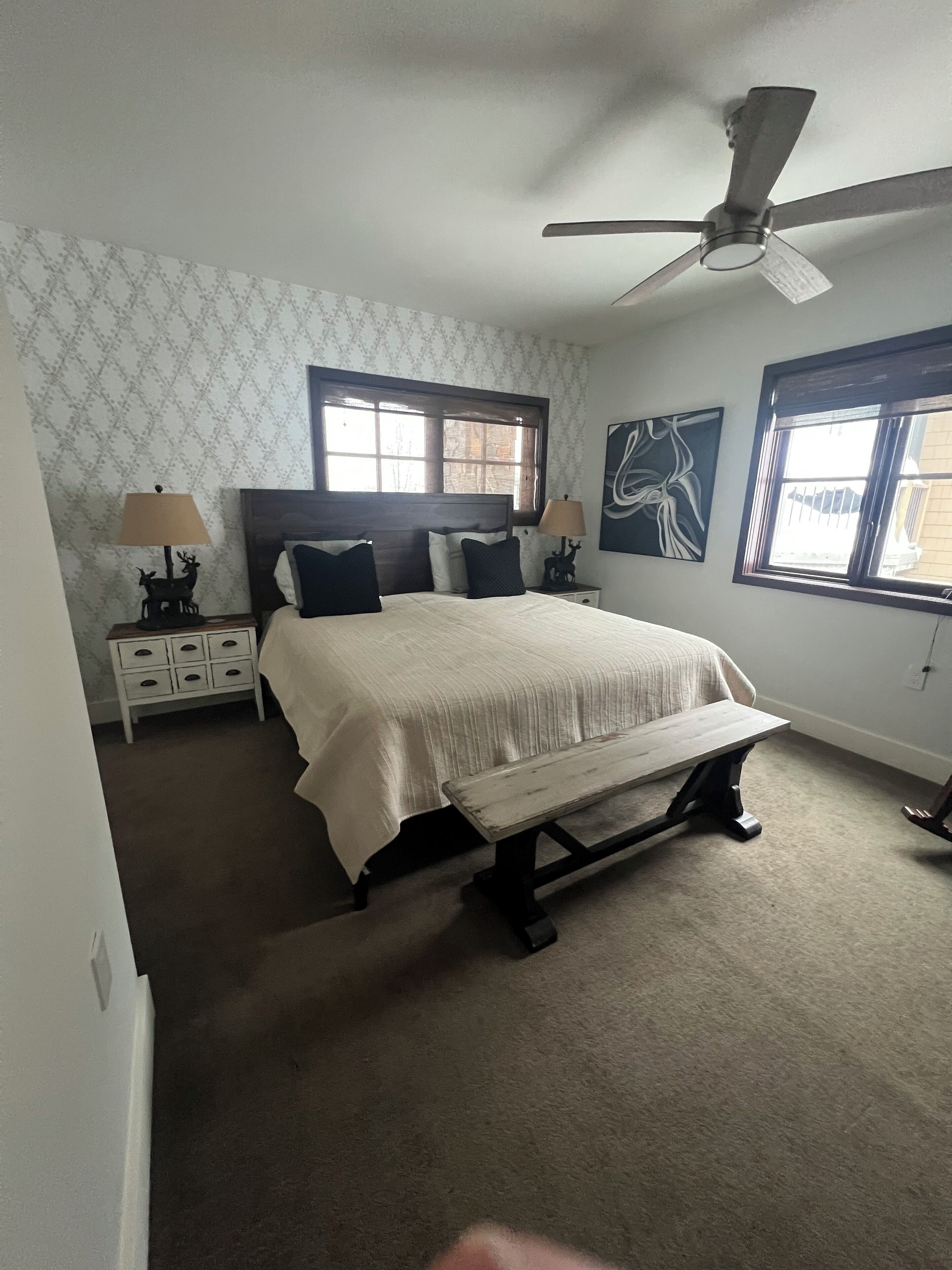 A bedroom with a bed, two nightstands, a bench, a ceiling fan, and two windows.