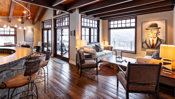A spacious room with a wooden ceiling, large windows, a bar area with stools, and a cozy seating arrangement. - Ski-in/Ski-out, In the Village, 7 King Bedrooms, 2 Bedrooms w/ 2 XL twins (Sandpoint)