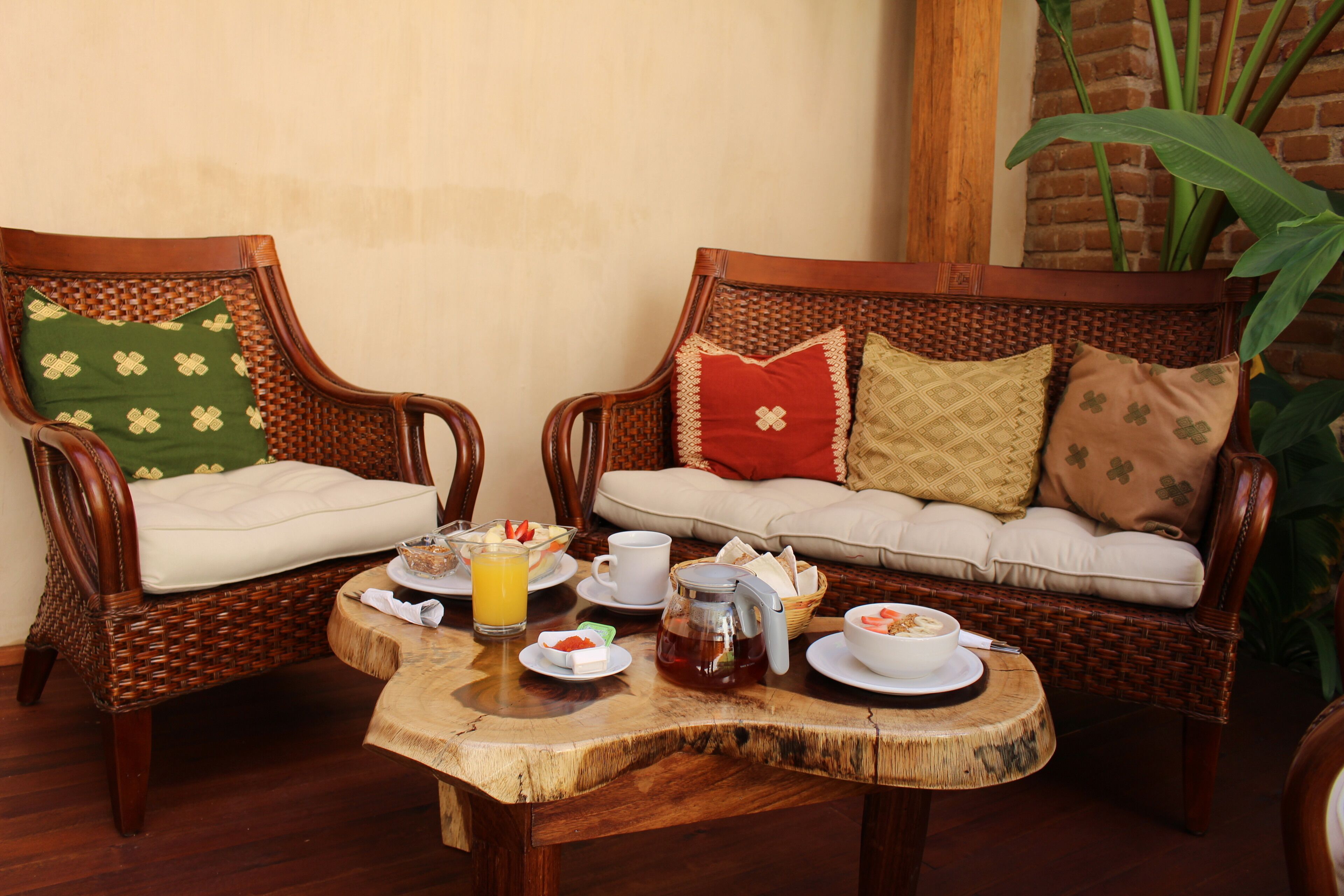 Two wicker chairs with cushions, a small table with breakfast items, and a brick wall with a plant.