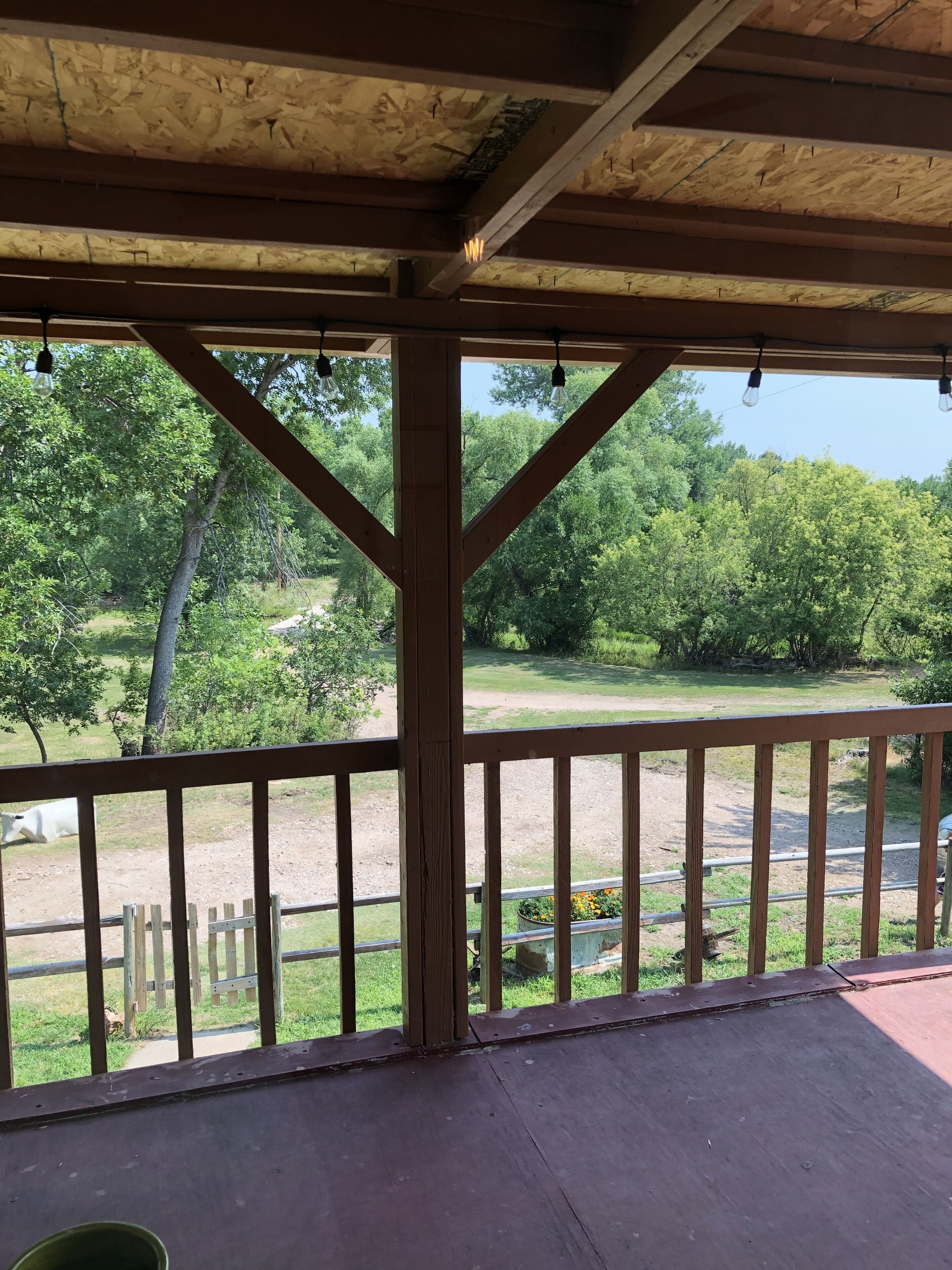 A covered outdoor area with a wooden deck and railings, overlooking a grassy field and trees.