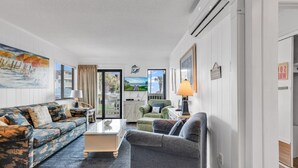 Una sala moderna con sofá, sillón y mesa de centro. Hay un cuadro en la pared, una lámpara y vistas al exterior a través de un ventanal.