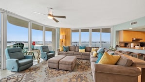 A spacious living room with a large sectional sofa, two armchairs, and a coffee table. A ceiling fan and a view of the city are visible. - Elegant Media Room Unit with Sparkling Bay Views! Complimentary Boat Slip! (Orange Beach)