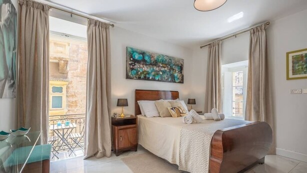 A bedroom with a bed, nightstands, a painting, and a view of the outside.