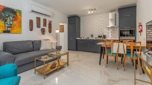 A compact living space with a grey sofa, a coffee table, a dining area with wooden chairs, and a kitchen with grey cabinets and a white tiled backsplash.