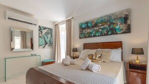 A modern hotel room with a large bed, a wooden headboard, a large abstract painting, a mirror, and a bedside table with a lamp.