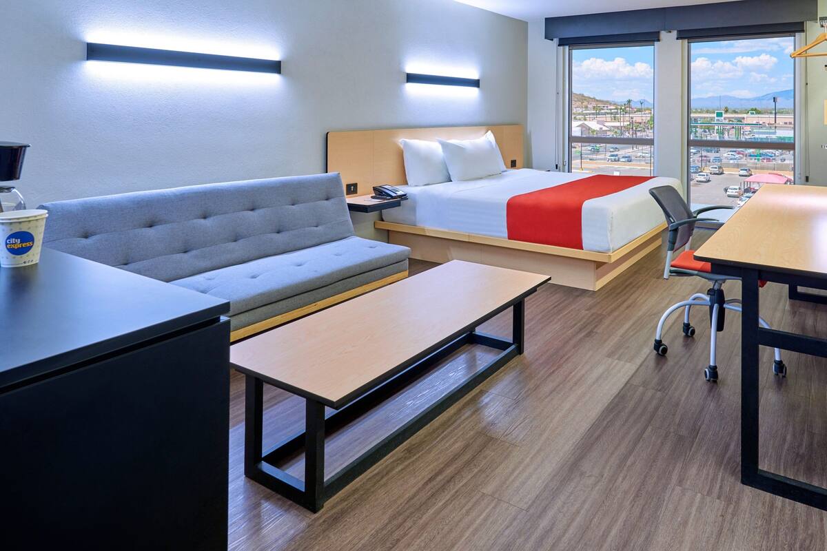 A hotel room with a bed, a sofa, a desk, and a view of the city through large windows.