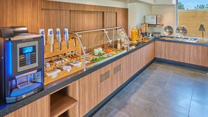 Una estación de desayuno autoservicio con una máquina de café, diferentes opciones de comida y un dispensador de bebidas.