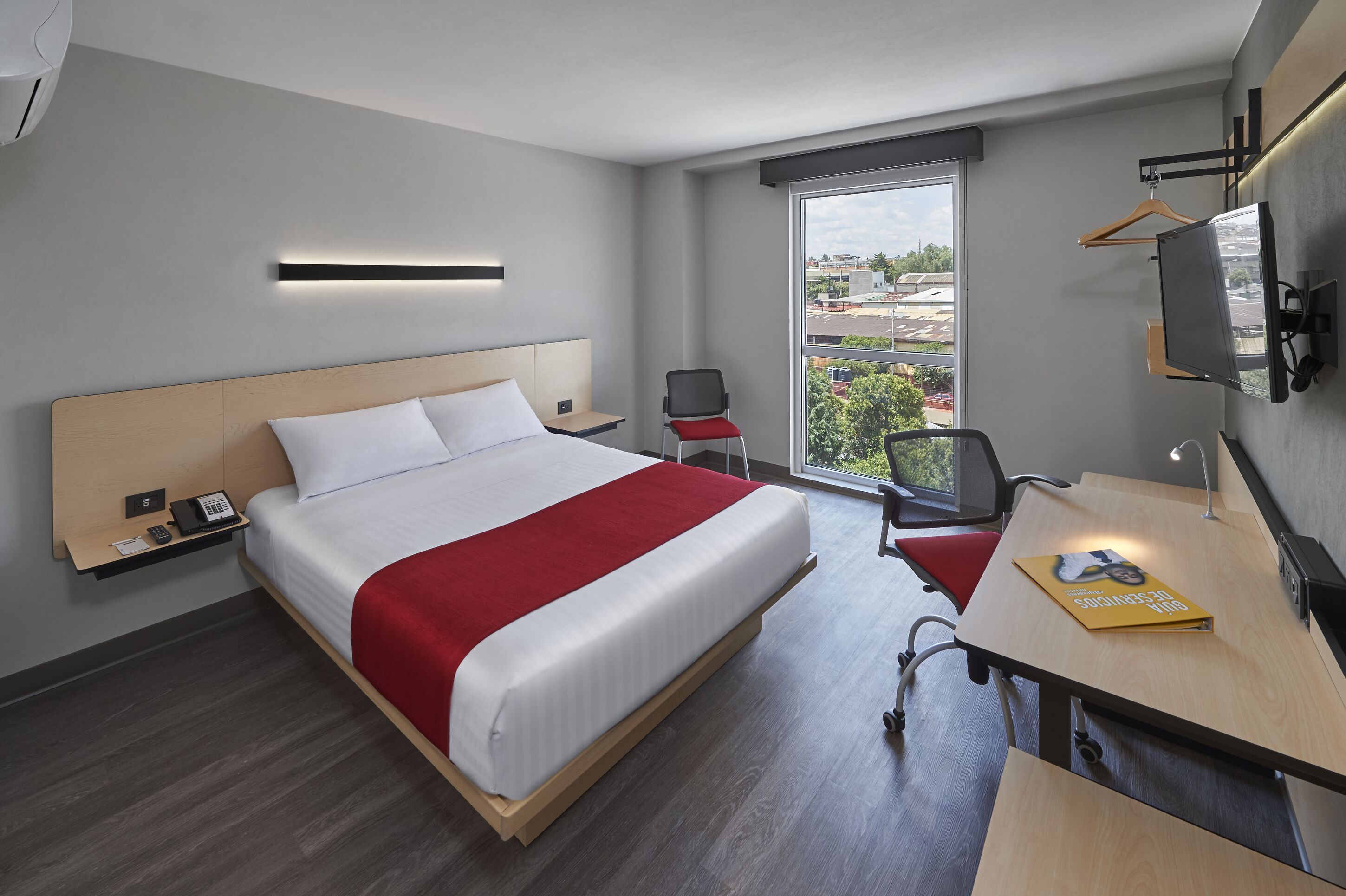 A hotel room with a large bed, a desk with a chair, a TV, and a window with a view of buildings.
